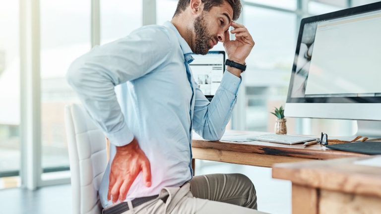 A man sitting at a desk appears to be experiencing back pain while working on a computer.